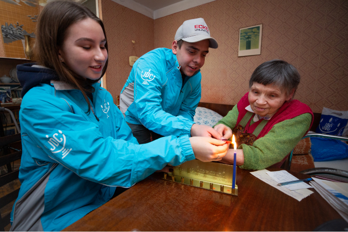 Two young JDC volunteers in the former Soviet Union light Channukah candles with an elderly client in her living room.