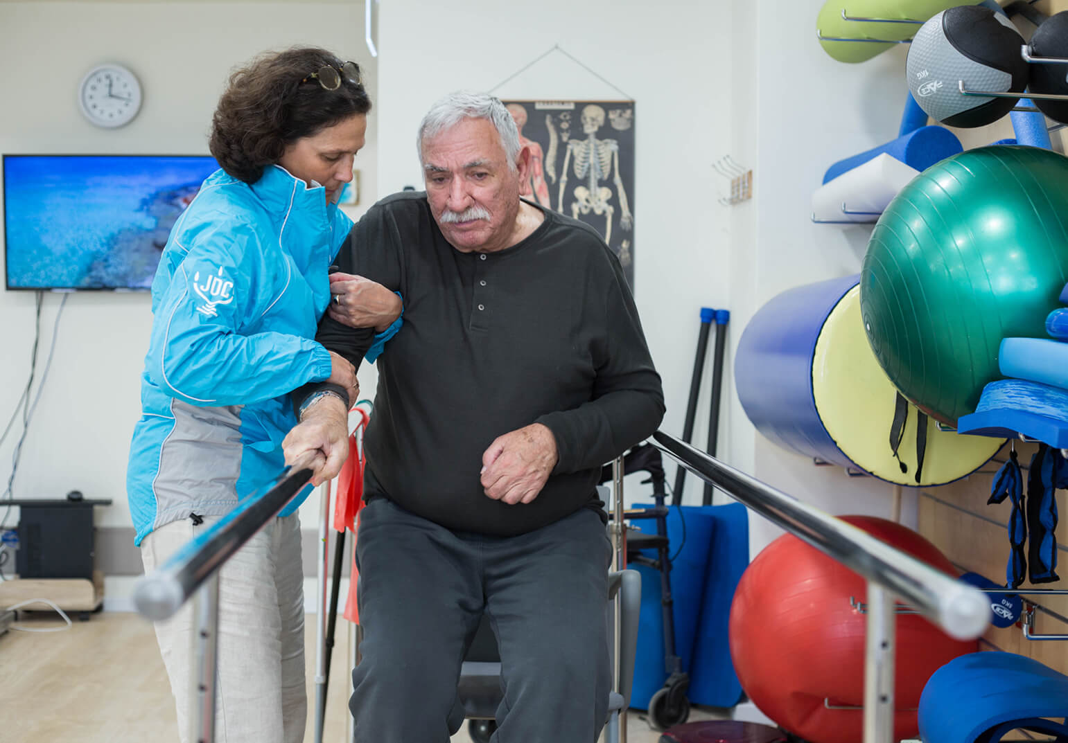 A woman in a JDC blue jacket lifts up an elderly Jewish man in a black shirt and sweatpants during a physical therapy class in Israel.