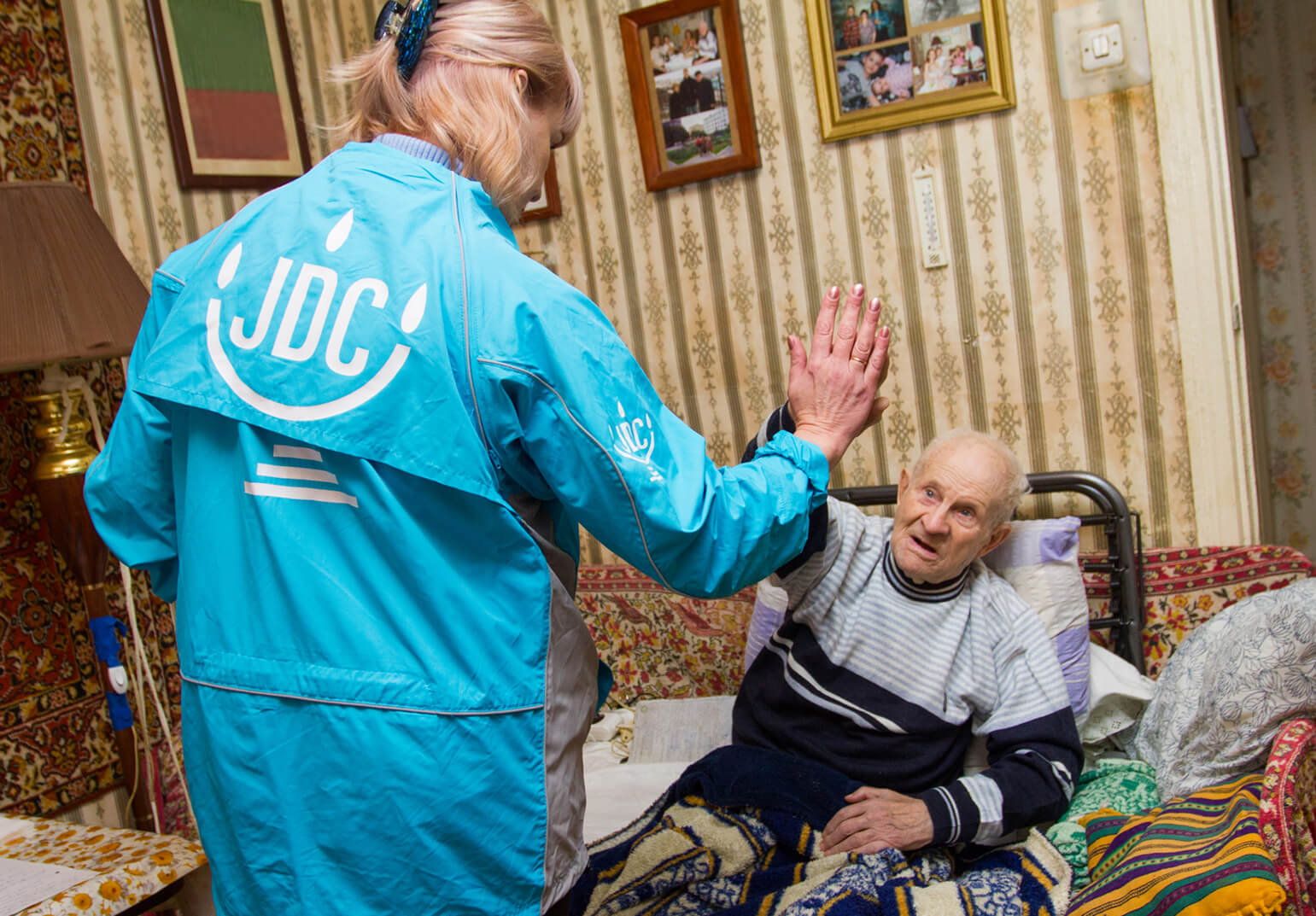 A blond woman in a JDC blue jacket high-fives a smiling elderly Jewish man sitting on a couch in Ukraine.