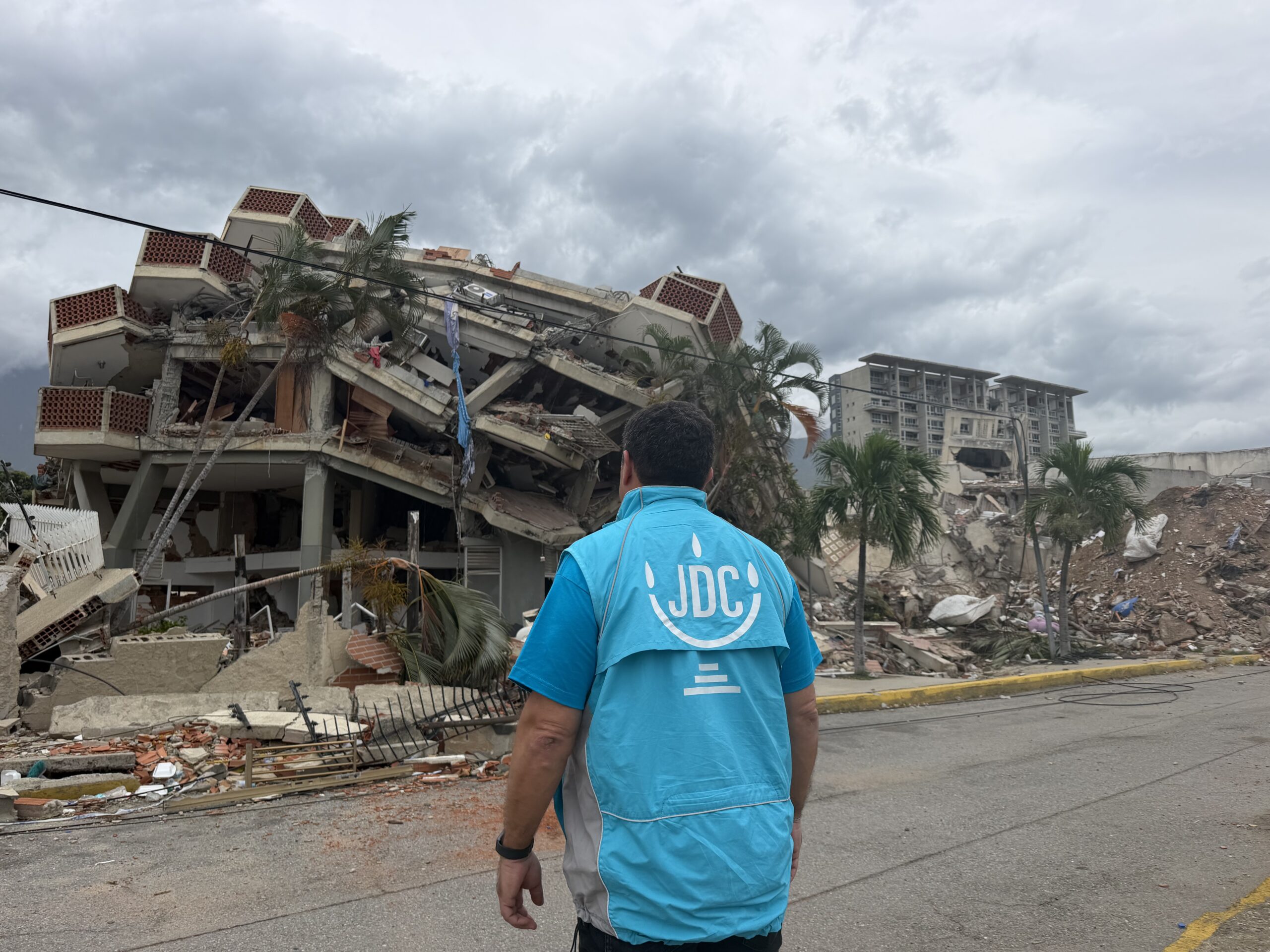 Person wearing a bright blue jacket stands in front of a collapsed building with rubble and debris under a cloudy sky.